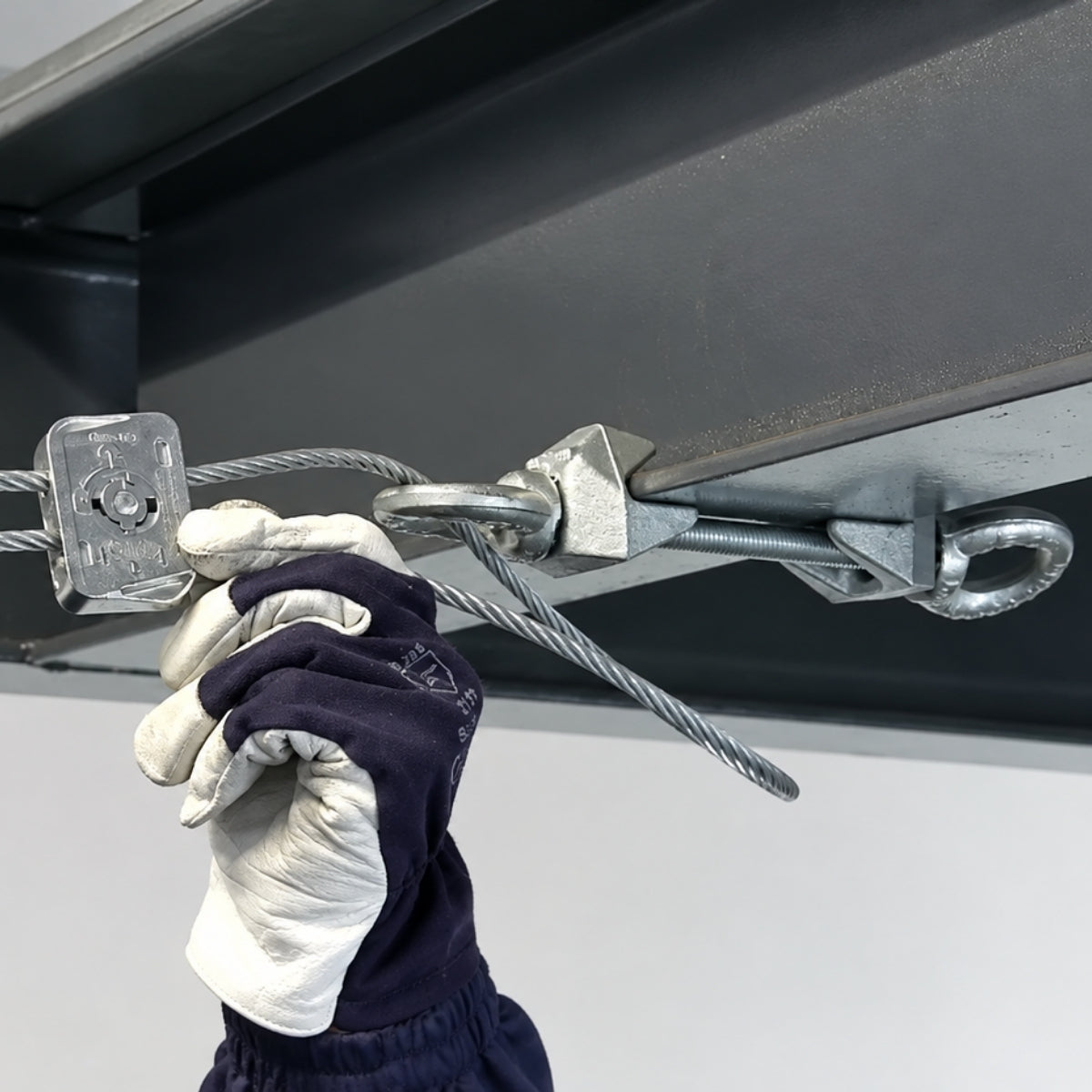 A gloved hand fitting a Gripple Dynamic to overhead wires under a ceiling.