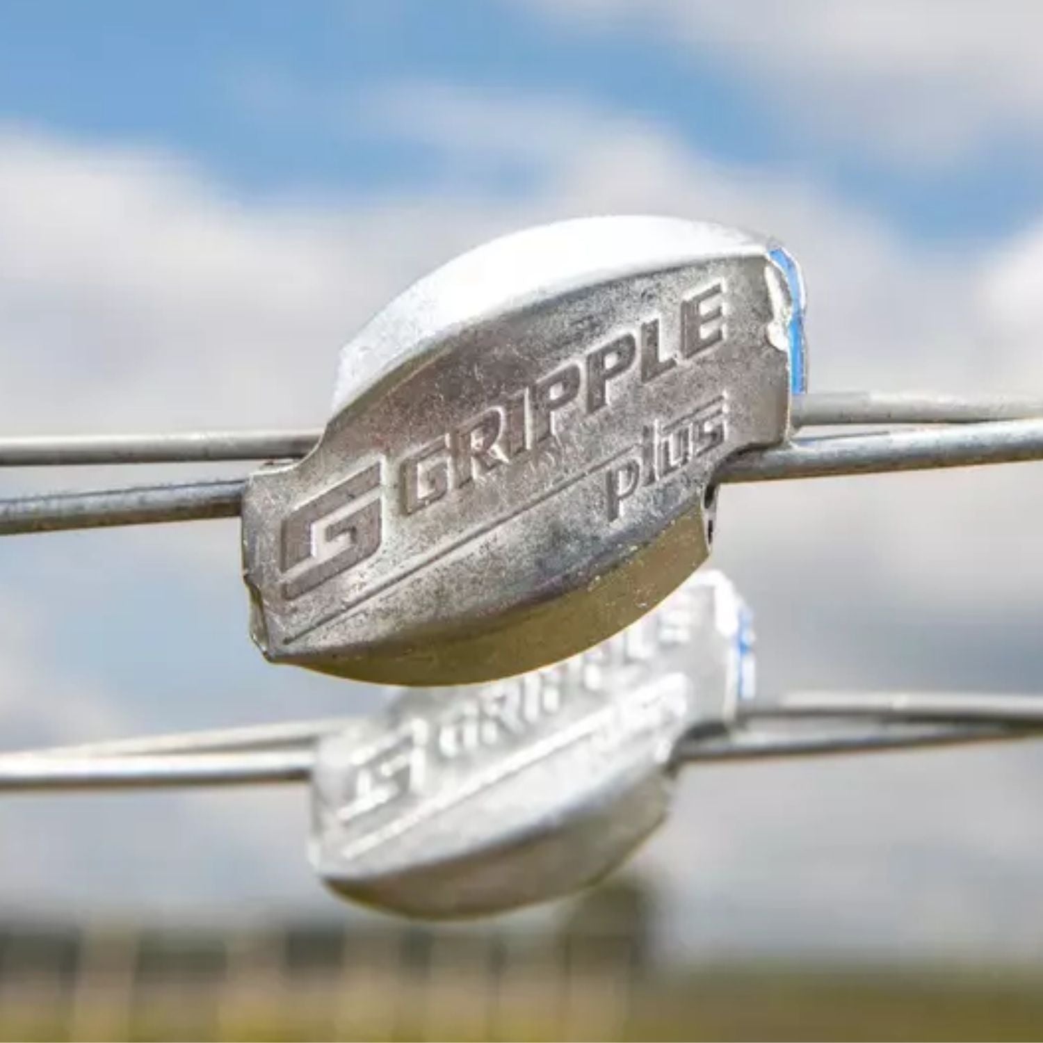 Close-up of a Gripple Plus wire connector installed on a high-tensile metal fence wire line under a cloudy sky.