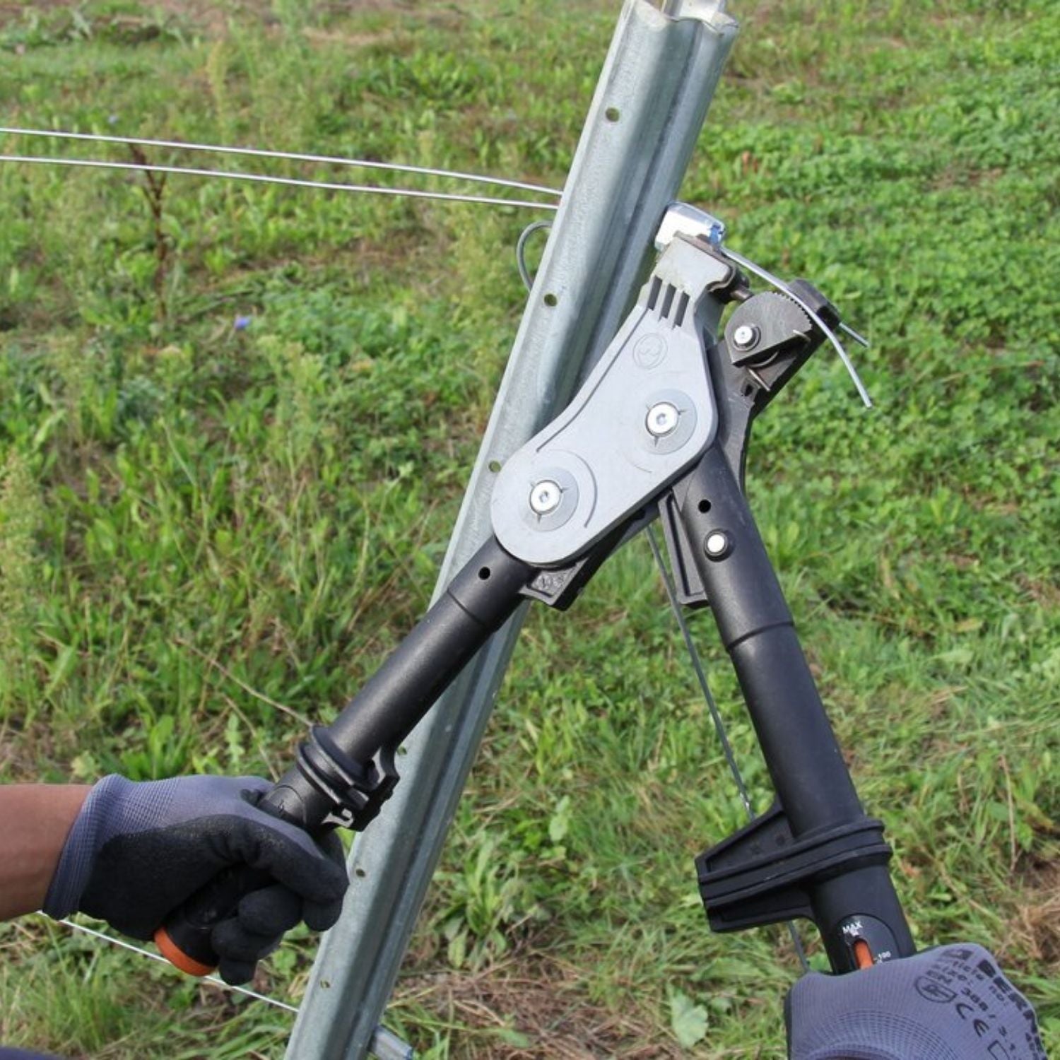 An installer using a Gripple torq tensioning tool to tighten the wire line directly against a metal trellis post in a vineyard field.